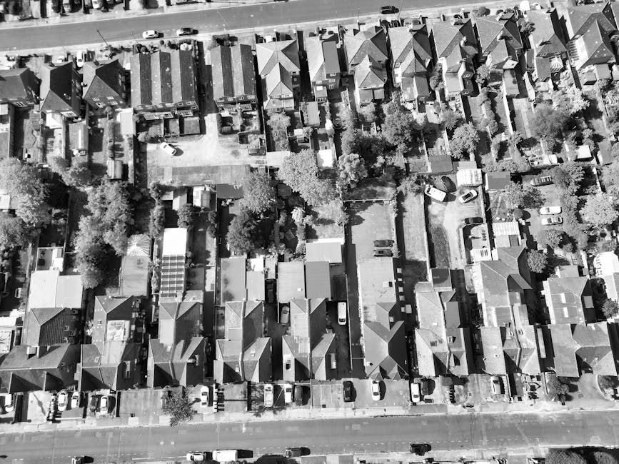 An aerial black-and-white photograph of a residential suburban area showing a dense row of houses with pitched roofs, tree-lined streets, and small gardens. The gardens feature neatly trimmed hedges, 